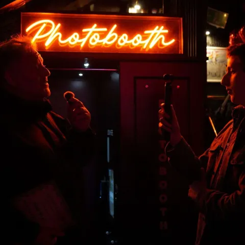 A woman smiles while talking to a man in a dimly lit bar with neon signage in the background.