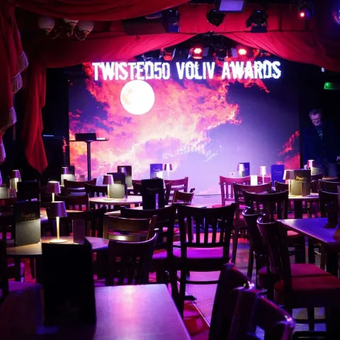 A dimly lit, empty bar or cabaret with scattered tables and chairs, red and purple lighting, framed pictures on dark walls, and small lamps on some tables.