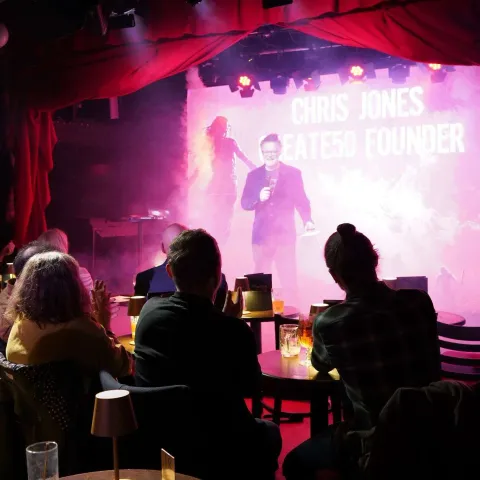 Three men stand together in a dimly lit bar, smiling and holding drinks, surrounded by other patrons and colorful lighting.