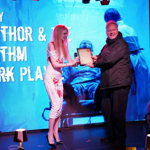 A woman in a white dress hands a plaque to a man on a stage, with a large blue screen displaying text in the background.
