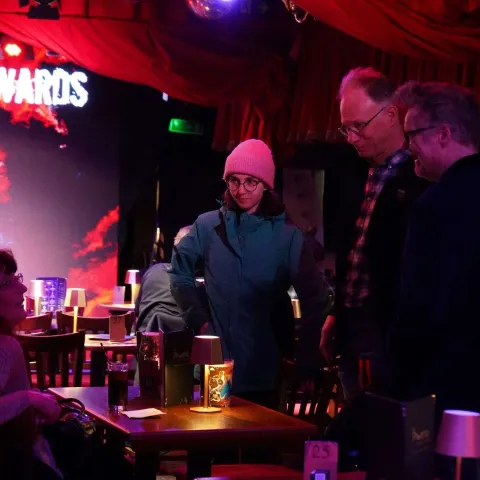 Two women sit at a table in a dimly lit bar, engaged in conversation. Other patrons are visible in the background under red lighting.