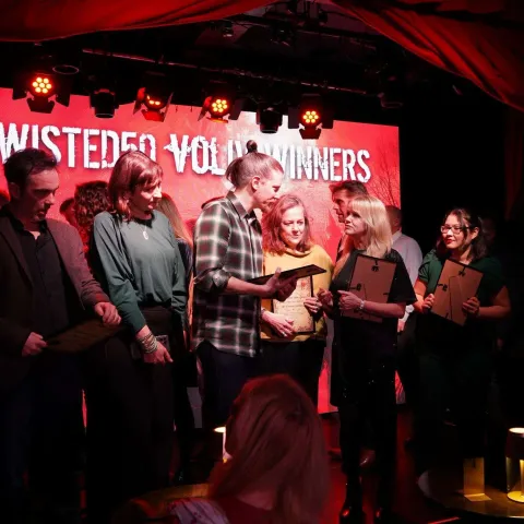 A group of people on stage receive awards, standing in front of a red screen that reads "TWISTED50 VOL 3 WINNERS" under stage lighting.