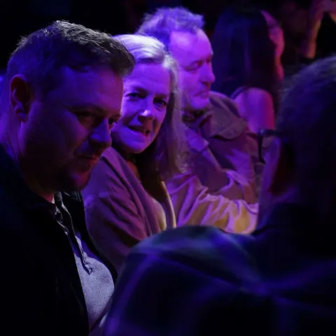 A woman smiles while talking to a man in a dimly lit bar with neon signage in the background.