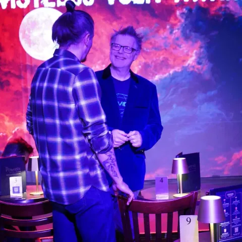 A woman smiles while talking to a man in a dimly lit bar with neon signage in the background.