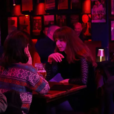 Two women sit at a table in a dimly lit bar, engaged in conversation. Other patrons are visible in the background under red lighting.