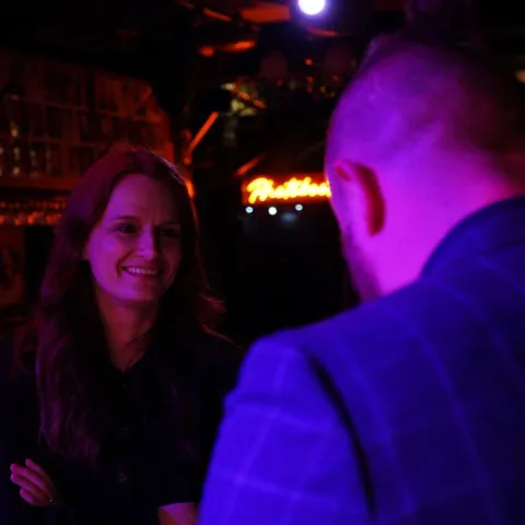 A woman smiles while talking to a man in a dimly lit bar with neon signage in the background.