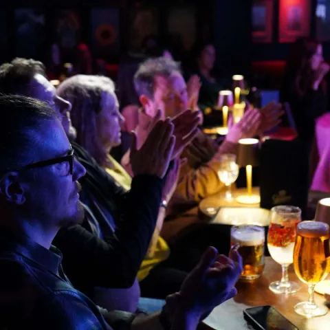Three men stand together in a dimly lit bar, smiling and holding drinks, surrounded by other patrons and colorful lighting.