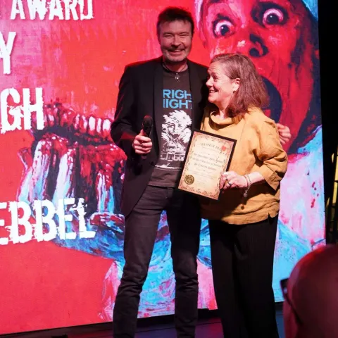 A person in a red beanie and plaid shirt holds a microphone and a plaque while standing on stage in front of a screen displaying text.