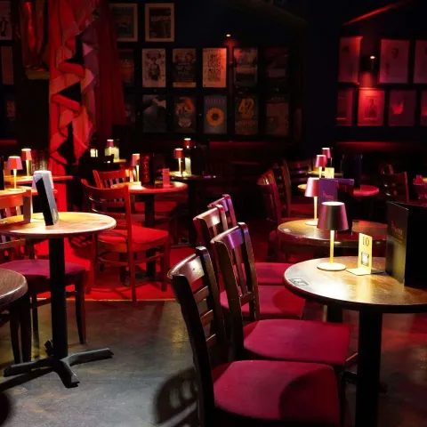 Two women sit at a table in a dimly lit bar, engaged in conversation. Other patrons are visible in the background under red lighting.