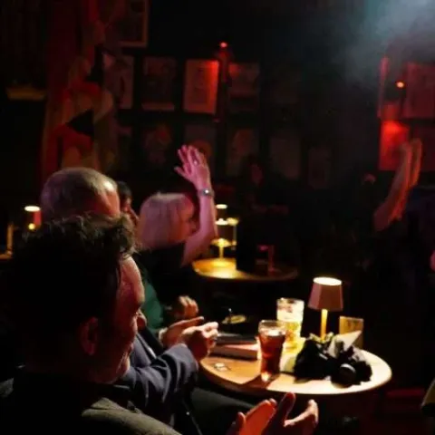Three people stand talking at a small table in a dimly-lit bar with red curtains and a "Twisted Vision Awards" sign visible in the background.