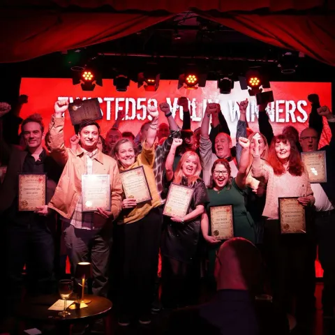 People are seated at small round tables in a dimly lit venue, watching a presentation on stage with a screen displaying "TWISTED50 VOL IV AWARDS.
