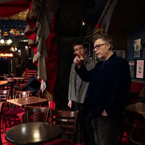Two women sit at a table in a dimly lit bar, engaged in conversation. Other patrons are visible in the background under red lighting.