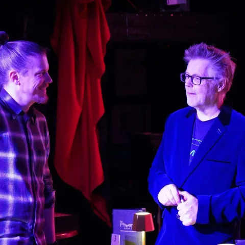 Three people stand talking at a small table in a dimly-lit bar with red curtains and a "Twisted Vision Awards" sign visible in the background.