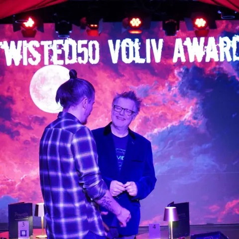 Three people stand talking at a small table in a dimly-lit bar with red curtains and a "Twisted Vision Awards" sign visible in the background.