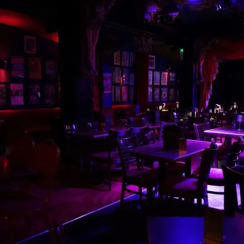 A dimly lit, empty bar or cabaret with scattered tables and chairs, red and purple lighting, framed pictures on dark walls, and small lamps on some tables.