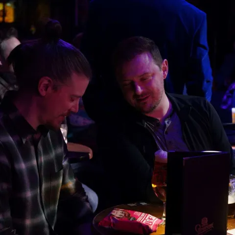 Three people stand talking at a small table in a dimly-lit bar with red curtains and a "Twisted Vision Awards" sign visible in the background.