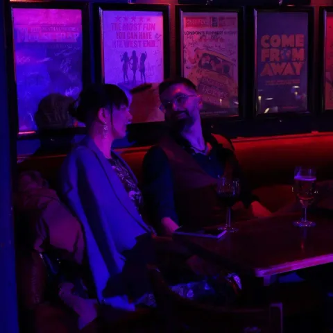 Three people stand talking at a small table in a dimly-lit bar with red curtains and a "Twisted Vision Awards" sign visible in the background.
