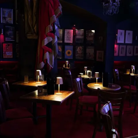 Dimly lit cabaret-style venue with small tables, red chairs, table lamps, red curtains, and framed posters on the dark walls.