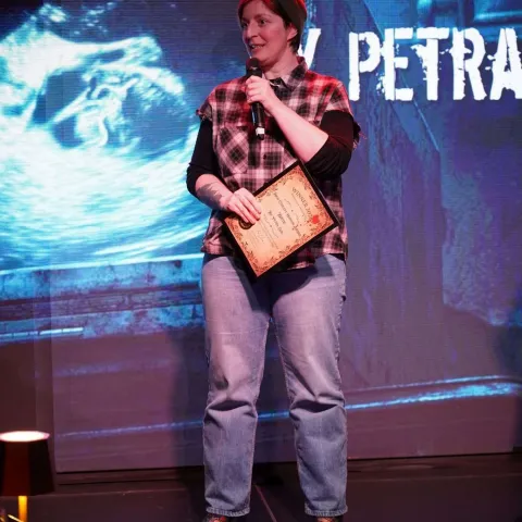A person in a red beanie and plaid shirt holds a microphone and a plaque while standing on stage in front of a screen displaying text.