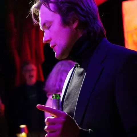 A woman smiles while talking to a man in a dimly lit bar with neon signage in the background.