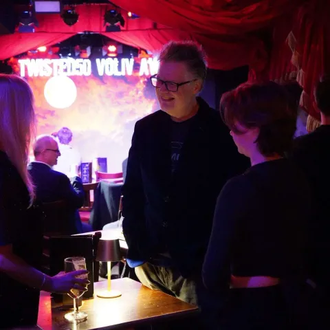 Three people stand talking at a small table in a dimly-lit bar with red curtains and a "Twisted Vision Awards" sign visible in the background.