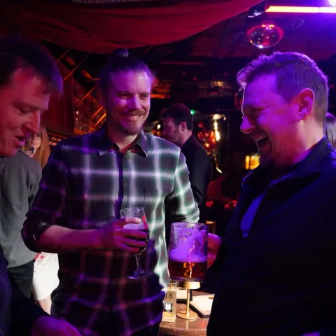 Three men stand together in a dimly lit bar, smiling and holding drinks, surrounded by other patrons and colorful lighting.