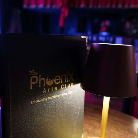 Dimly lit cabaret-style venue with small tables, red chairs, table lamps, red curtains, and framed posters on the dark walls.