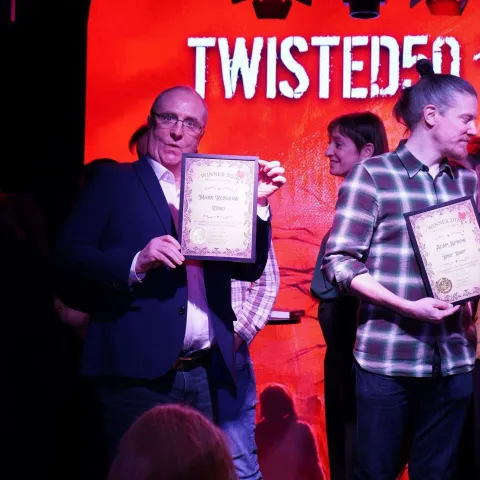 People are seated at small round tables in a dimly lit venue, watching a presentation on stage with a screen displaying "TWISTED50 VOL IV AWARDS.