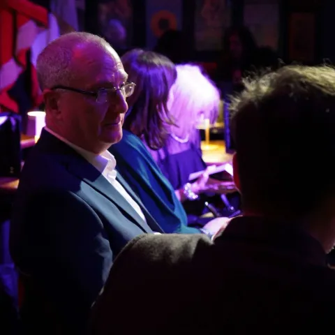 Two women sit at a table in a dimly lit bar, engaged in conversation. Other patrons are visible in the background under red lighting.