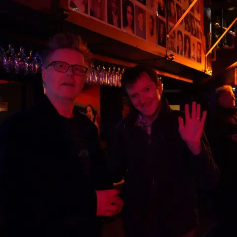 A woman smiles while talking to a man in a dimly lit bar with neon signage in the background.