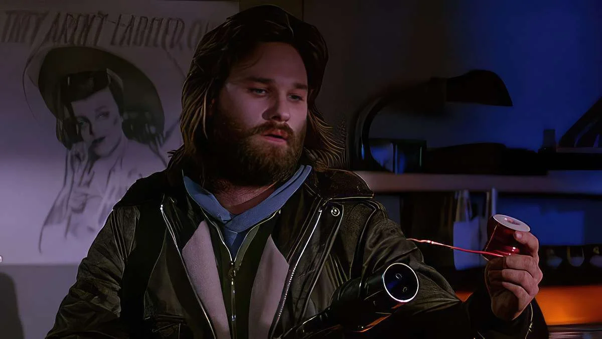 A man with long hair and a beard, wearing a jacket, holds a spool of wire in one hand while examining it. There is a poster of a woman in the background and the scene is dimly lit.