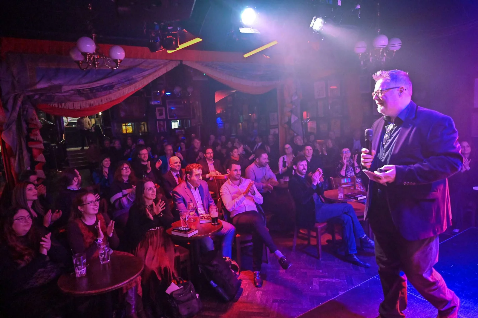 A man stands on stage holding a microphone, speaking to a seated audience in a dimly lit venue. Some audience members are clapping, and tables with drinks are visible.