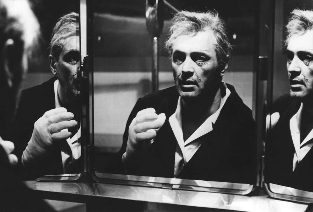 A man with disheveled hair and a worn expression looks at his reflection in a mirror, raising a gloved hand toward his face.