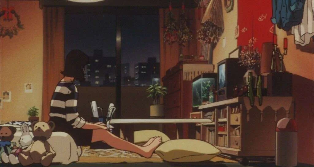 A person sits on the floor in a cozy, dimly lit room watching TV, surrounded by stuffed animals, pillows, and various decorations, with a cityscape visible through the window.