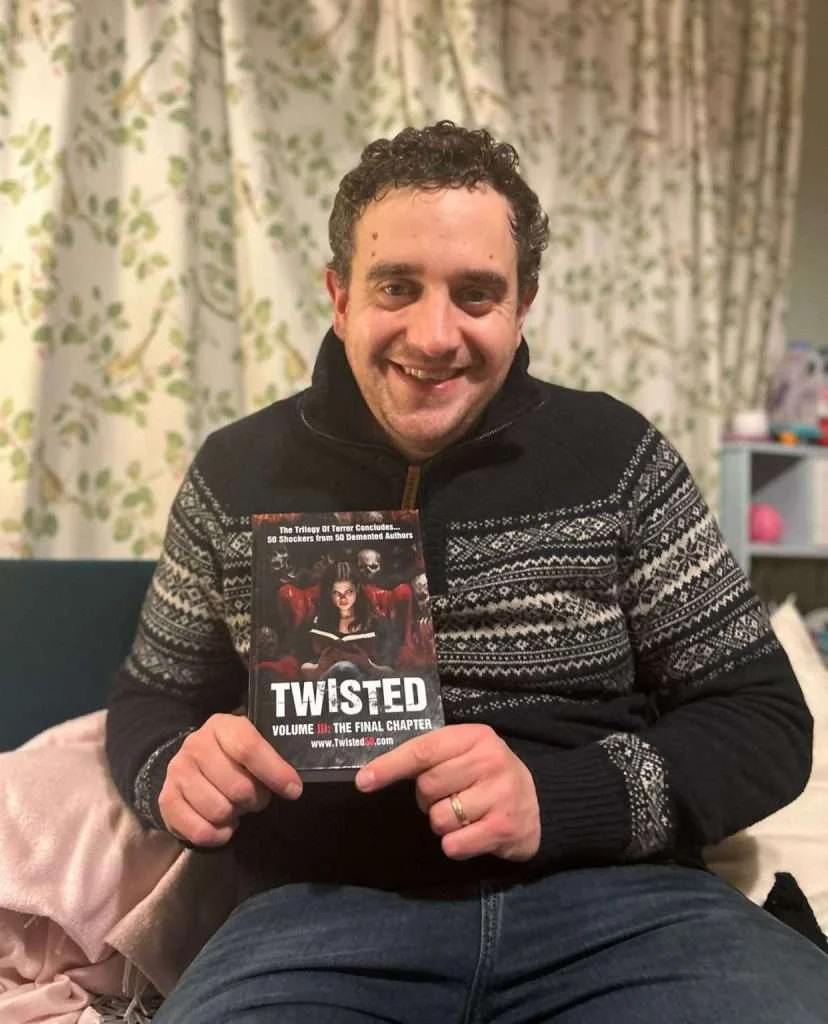 Man sitting on a couch, smiling and holding a book titled "Twisted: The Final Chapter." The background shows a floral curtain and household items.