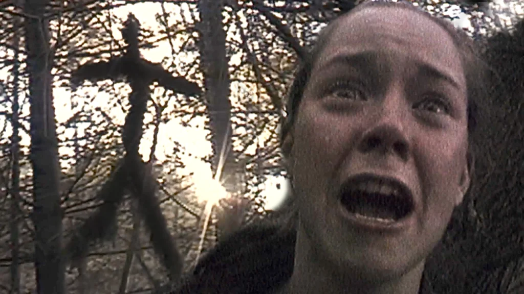 A distressed woman is screaming in a forest with sunlight behind her; a stick figure made of branches hangs from a tree in the background.