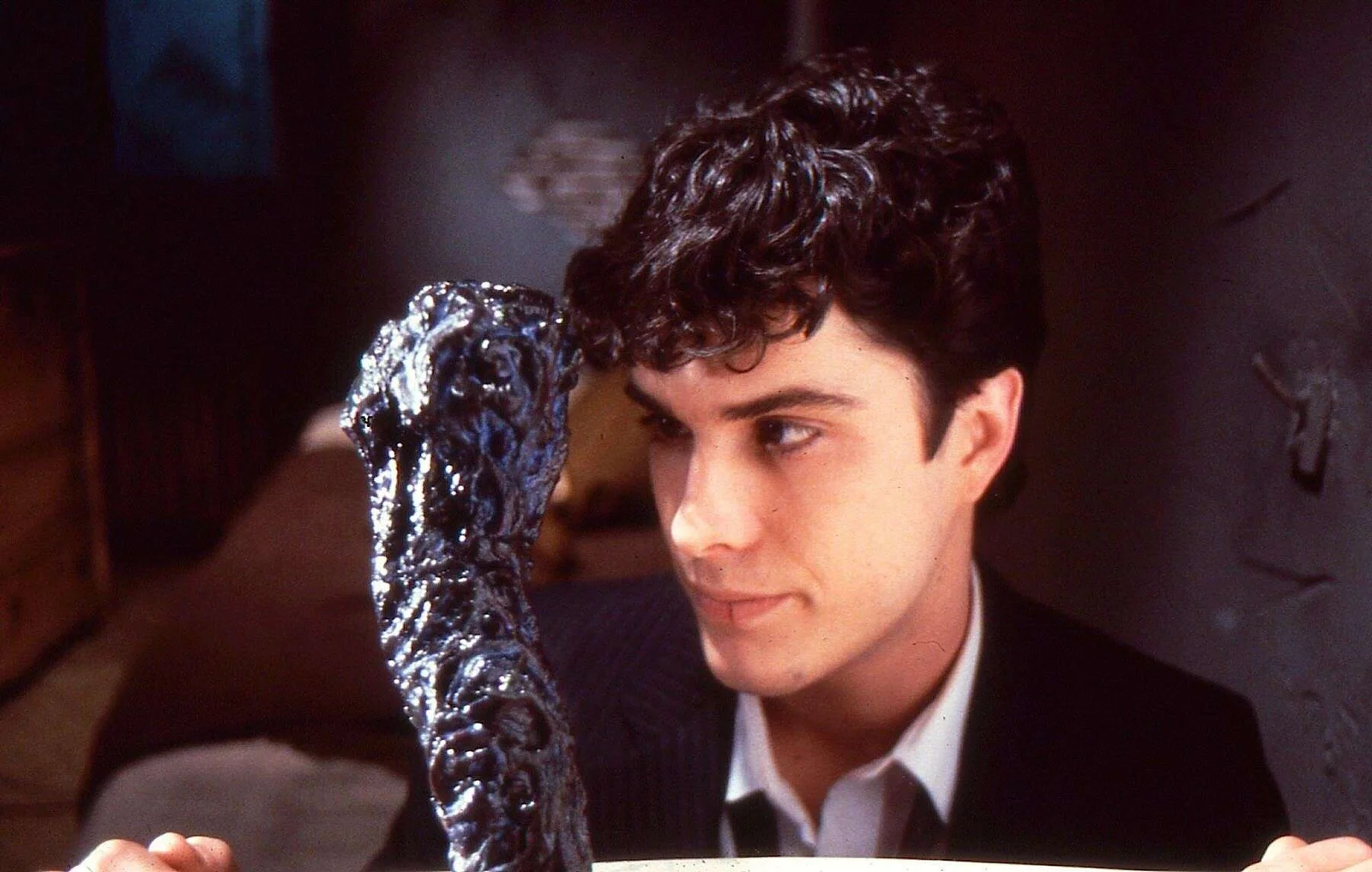 A young man in a suit looks closely at a dark, slimy, snake-like creature in a dimly lit room.